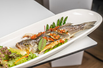 A beautifully presented whole grilled fish garnished with herbs, vegetables, red caviar, and served with fresh greens and lemon on a white plate