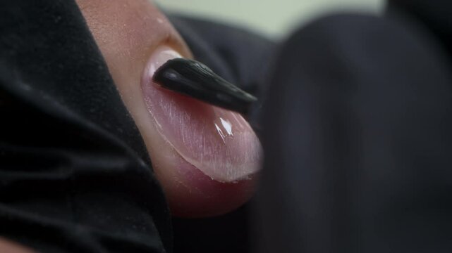 Close-up of nail technician wearing black gloves while applying clear gel to natural nail using small precision brush, creating clean and glossy base layer