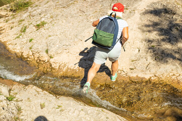 Active senior woman in shorts, red cap, and backpack crossing a shallow mountain stream during a...
