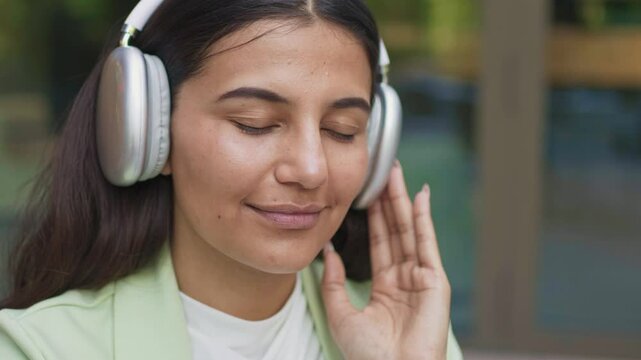 Charming woman with dark straight hair wearing headphones enjoying music with eyes closed. Slightly smiling. Feeling relaxed or immersed in sound. Touching earcup while listening outdoors.