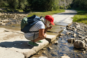 Elderly female hiker squatting and drinking clear water from a mountain stream. Wearing a red cap...