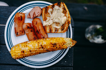 Grilled sausages, corn, and bread arranged on a white plate. A delicious plate of grilled food, perfect for a summer barbecue. Includes sausages, corn on the cob, and bread.