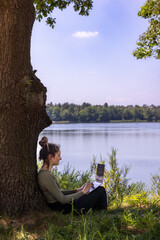 A young woman diligently working on her laptop by the serene and peaceful lakeside underneath a tree