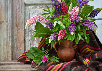 A bouquet of purple and pink lupine flowers in a clay jug on an old wooden background. Summer still life with flowers