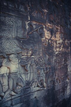Ancient stone wall with detailed bas-relief depicting warriors, elephants, and battle scenes. The carving shows movement and history, worn by time but rich in storytelling.