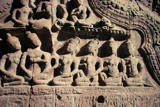 Detailed bas-relief carving of seated celestial figures on an ancient temple wall. The sandstone artwork shows expressive gestures and elaborate hair, illuminated by warm sunlight.