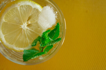 A glass of refreshing lemon drink with mint leaves and ice on a warm yellow background. A vibrant...