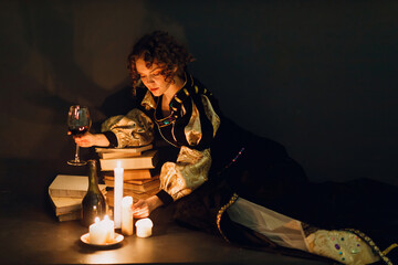 Young adult woman dressed in a medieval dress sitting near ancient books and candle light with...