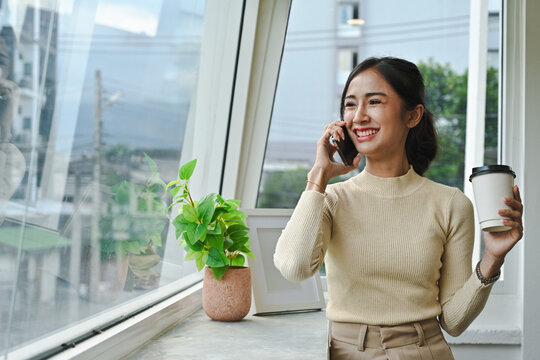 Confident businesswoman smiling while talking on the phone, holding a takeaway coffee cup in a bright, modern office environment