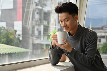 A young man in a gray sweater holding a white coffee mug, enjoying a peaceful moment by the window with soft natural light