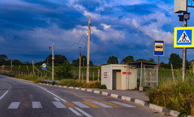A photograph of a rural bus stop against the backdrop of vineyards in Crimea. A paved road with...