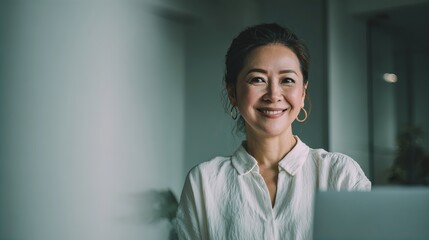 A smiling woman with dark hair pulled back, wearing a white blouse and gold hoop earrings, sits in a softly lit indoor space, exuding warmth and confidence.