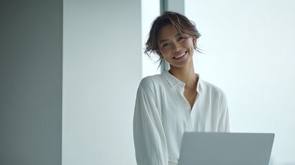 A smiling woman in a white blouse stands with a laptop, exuding confidence and professionalism in a bright, modern office setting.