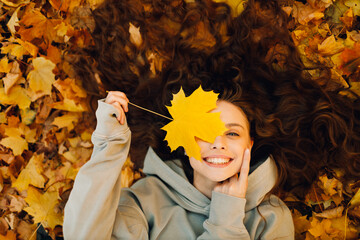 Smiling young woman lies on the autumn foliage in the forest with the yellow leaves