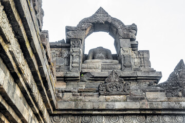A headless Buddha statue sits in a carved stone arch atop an ancient temple wall.
