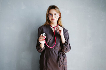 Portrait of female doctor in dark medical uniform using stethoscope and smiling at camera against gray background.