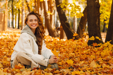 Smiling young woman sits in the autumn foliage on the ground of the forest with the yellow leaves
