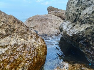 Water flows between large, textured rocks, leading to the ocean under a cloudy sky.