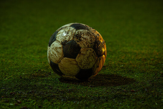 Muddy soccer ball lying on wet green grass field in evening stadium lighting. Concept of professional football training and fitness