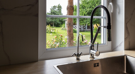 Kitchen sink with modern faucet, view through window to blooming garden, green trees, summer flowers, backyard landscape, cozy rural lifestyle, sustainable home, interior detail, daylight