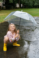 Kid playing in the rain. Little cheerful girl with a transparent umbrella and rubber boots sat in a...