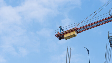 Detail of counterweight on red tower crane at construction site. Visible steel rebars and partly cloudy sky.