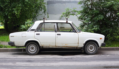 An old, rusty white car is parked near a lawn, Badayeva Street, Saint Petersburg, Russia, July 07, 2025 