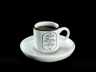 Dark coffee being poured into a white ceramic mug with a vintage Paris design, placed on a matching saucer against a black fabric background