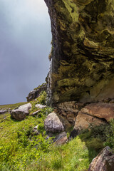 A small waterfall falling down a large sandstone overhang forming a vast cave, with sunlight glistering on the falling water with dark grey storm clouds in the background