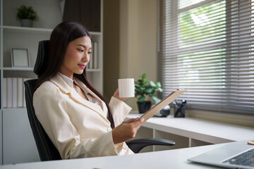 Confident businesswoman holding coffee cup and analyzing financial data in a bright modern office.
