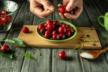 Hands meticulously remove stems from ripe cherries in a cozy kitchen. A vibrant bowl of cherries is...