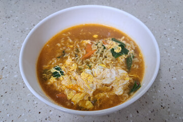 A bowl of ramen noodles with vegetables and eggs. The bowl is white and the noodles are orange