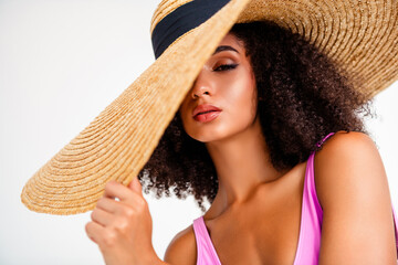 Stylish young woman in chic summer attire posing with a statement straw hat against a minimal white...