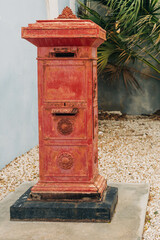 Vintage red mail box positioned in a serene outdoor setting with lush greenery nearby and gravel...