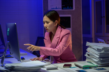 Stressed Asian businesswoman pointing at computer screen working late at night in office