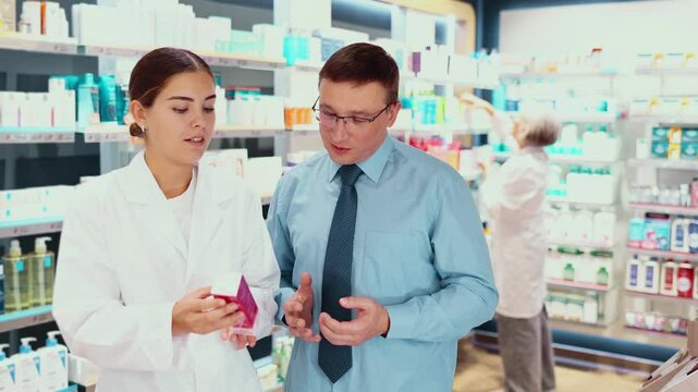 Polite young female pharmacist consulting middle-aged man costumer about care product in box in chemist's shop. High quality 4k footage