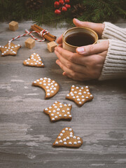 Christmas atmospheric set. Female hands in a sweater with coffee mug among tradition gingerbreads and holidays decoration on a wooden background, vertical image.