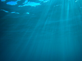 Dark blue ocean surface seen from underwater. Abstract waves underwater and rays of sunlight shining through, Sun light rays undersea deep, Underwater background with sea bottom, Mediterranean sea.