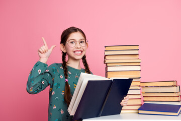 Smiling young girl gesturing a sign while holding book against a pink background surrounded by...