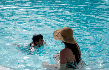 A woman is sitting in a pool with a child. The woman is wearing a straw hat and the child is...