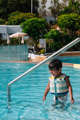 A young boy is in the water wearing a life jacket. He is standing in the pool and looking up at the camera