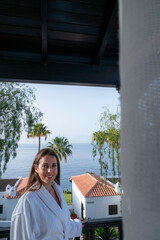 A woman is standing on a balcony overlooking the ocean. She is wearing a white robe and smiling. The scene is peaceful and relaxing, with the ocean and palm trees in the background