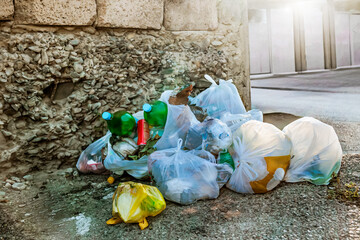 Pile of plastic garbage bags and bottles dumped near a rough stone wall on an urban street corner,...