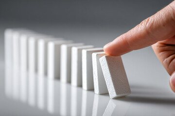 Finger gently pushes white domino piece creating systemic balance in sequence of falling dominoes on reflective surface