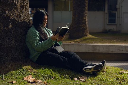 Woman in the park sitting on the grass with her phone and a book, calm and serene in the sun.