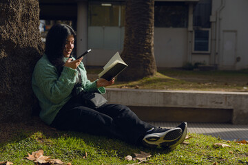 Young woman in the park sending a voice note on her phone while reading a book in the park