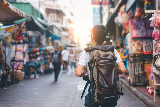 Rear view of a solo male traveler with a large backpack walking through a lively outdoor market street filled with colorful shops and people during sunset, representing travel, and urban adventure.