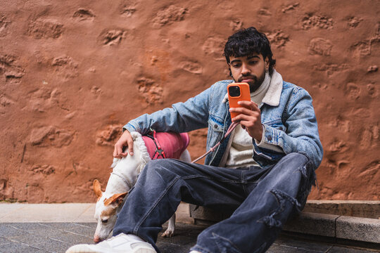 Stylish man using a smartphone and petting his dog while sitting on a step in front of a terracotta wall