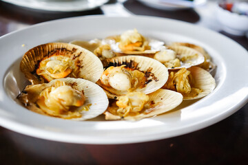 Grilled scallops in the shell with butter, a fresh seafood menu of a restaurant, served on a white plate on the wooden table with Thai seafood sauce.