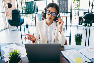 Businesswoman in a modern office talking through headphones during a video call on her laptop,...